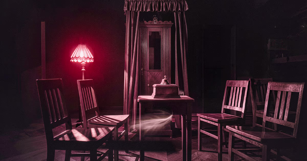 A dim parlour lit by a red lamp, with wooden chairs, a small bell box on a table and a cabinet. Illustration.