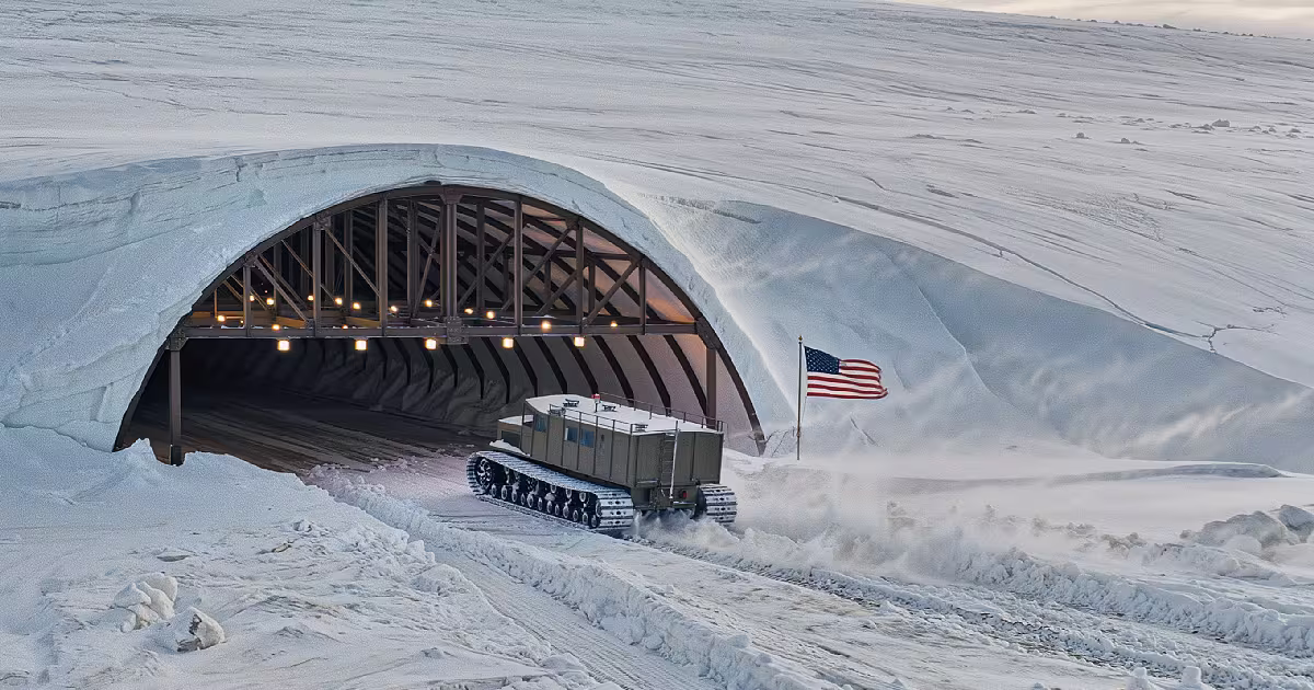 A tracked vehicle driving out of an arched trench entrance cut into the Greenland ice sheet, with a US flag in the snow beside it. Illustration.
